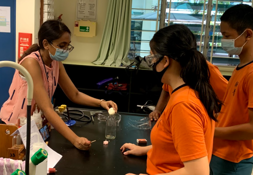 Teacher demonstrates a simple science experiment using a flask while students observe closely at a lab table.